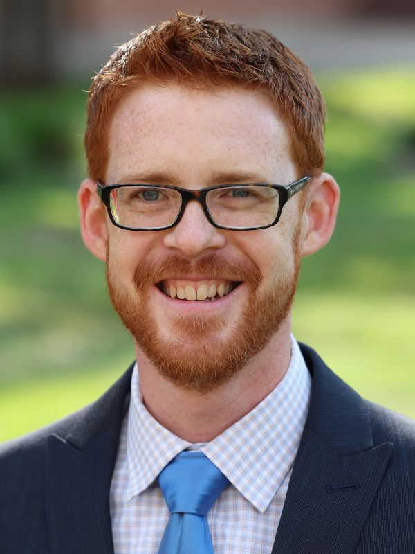 view of a smiling man with red hair and black glasses wearing a suit with a cornflower blue tie