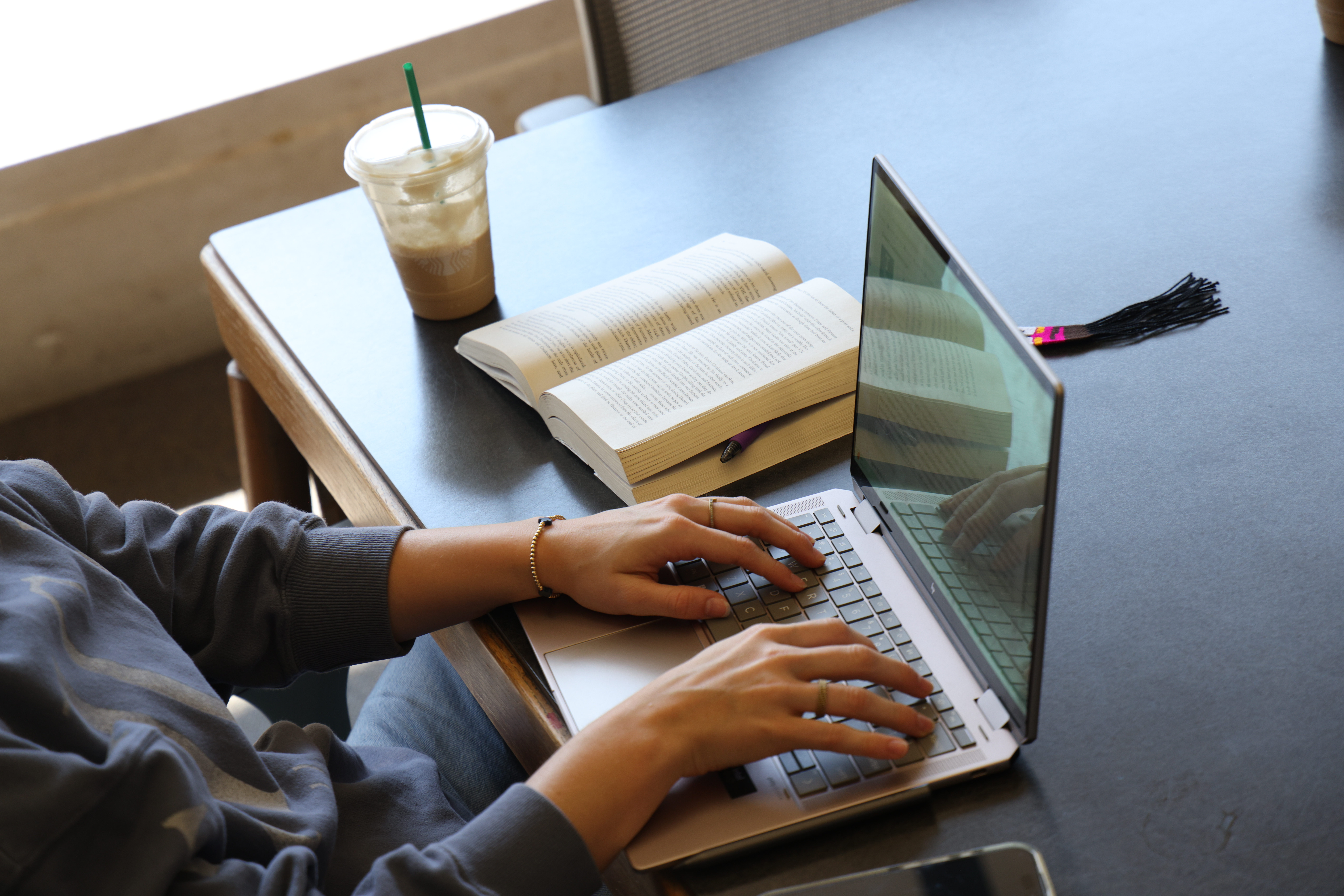 view of a computer with hands typing, with an open book and a coffee nearby