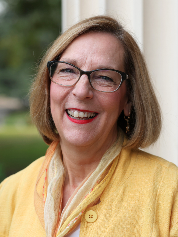view of a smiling woman in glasses with short blonde hair, wearing a golden shirt with a gold and yellow scarf